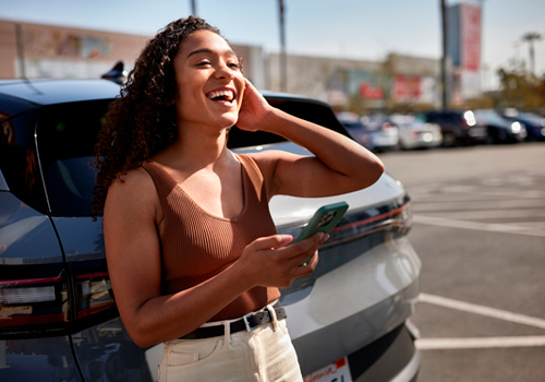 Jovem sorridente encostada ao carro com telemóvel na mão