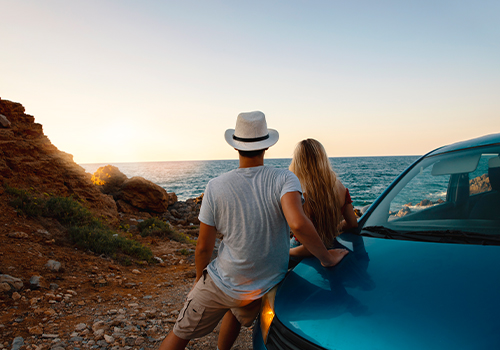 casal na berma da estrada a apreciar o mar encostados ao carro.